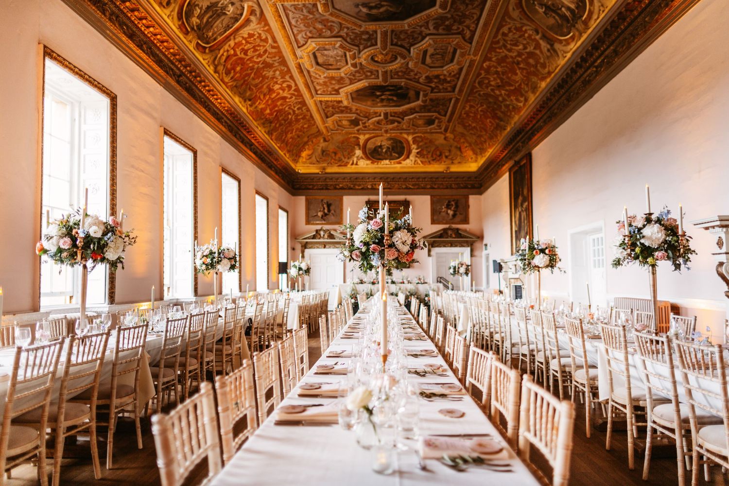 Long banquet table setup in grand hall with ornate painted ceiling and white chairs.