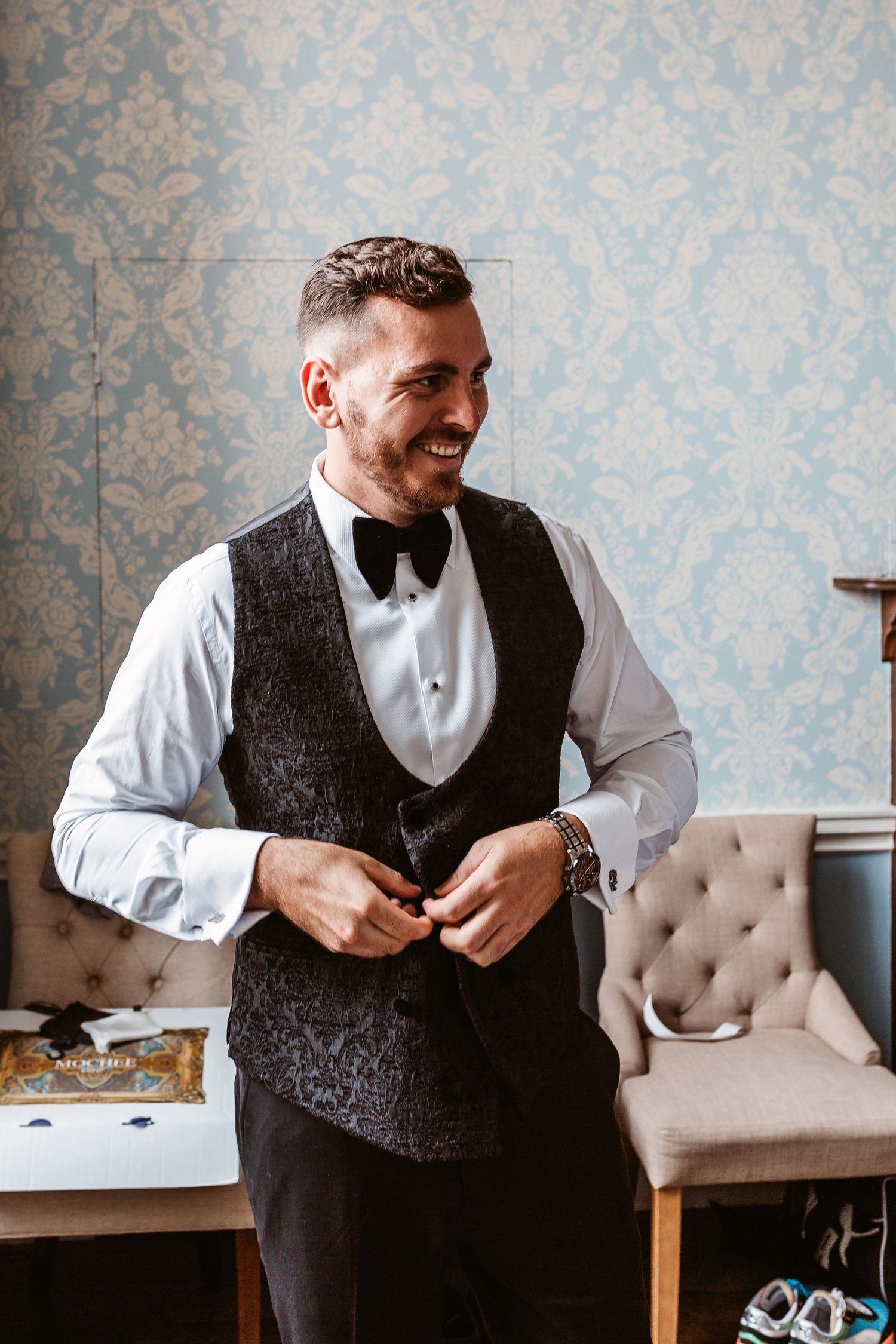 A person in formal attire with a dark vest and bowtie buttons their waistcoat against an elegant backdrop.