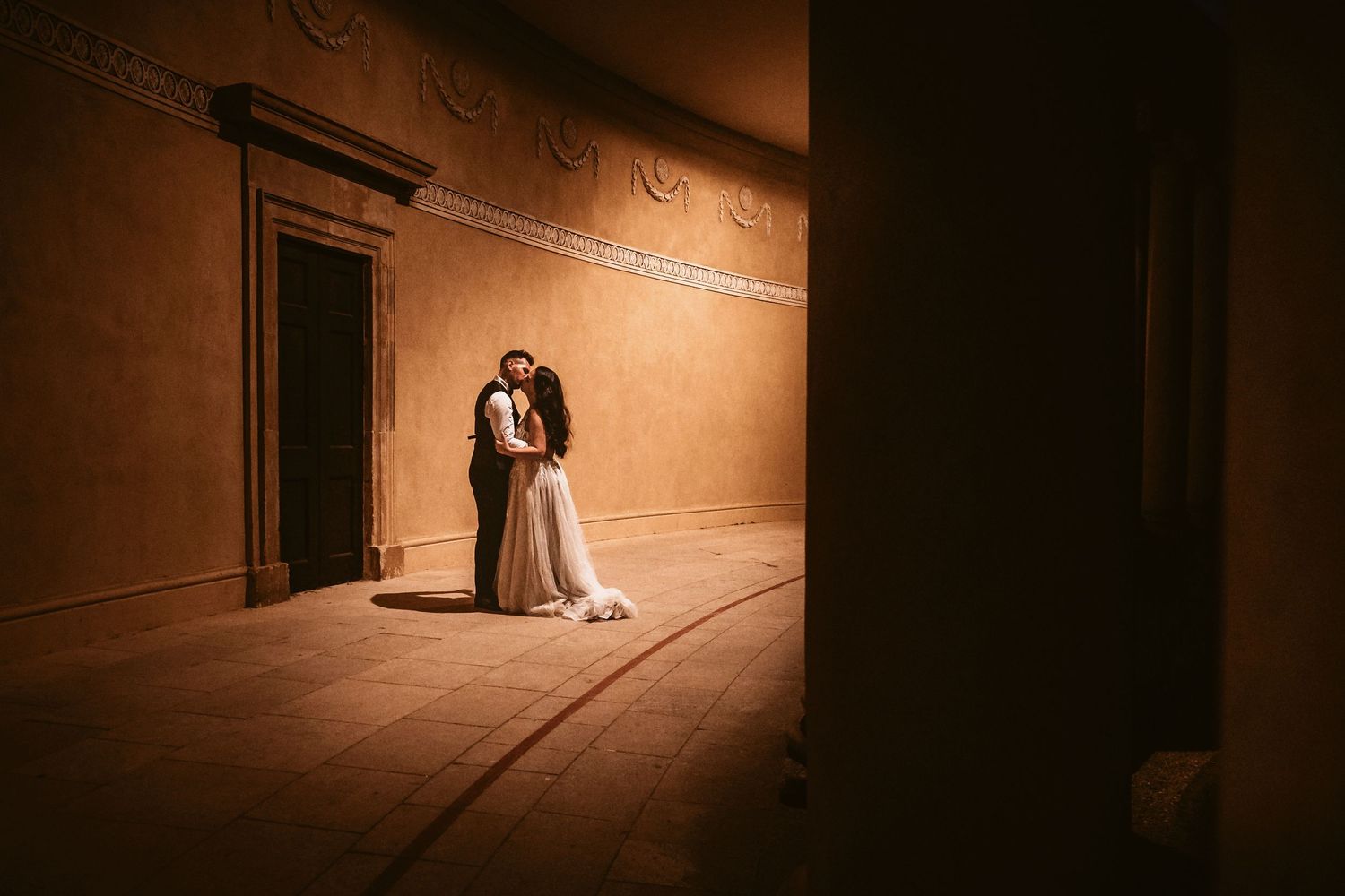 A romantic embrace in a curved hallway with dramatic lighting and architectural details.