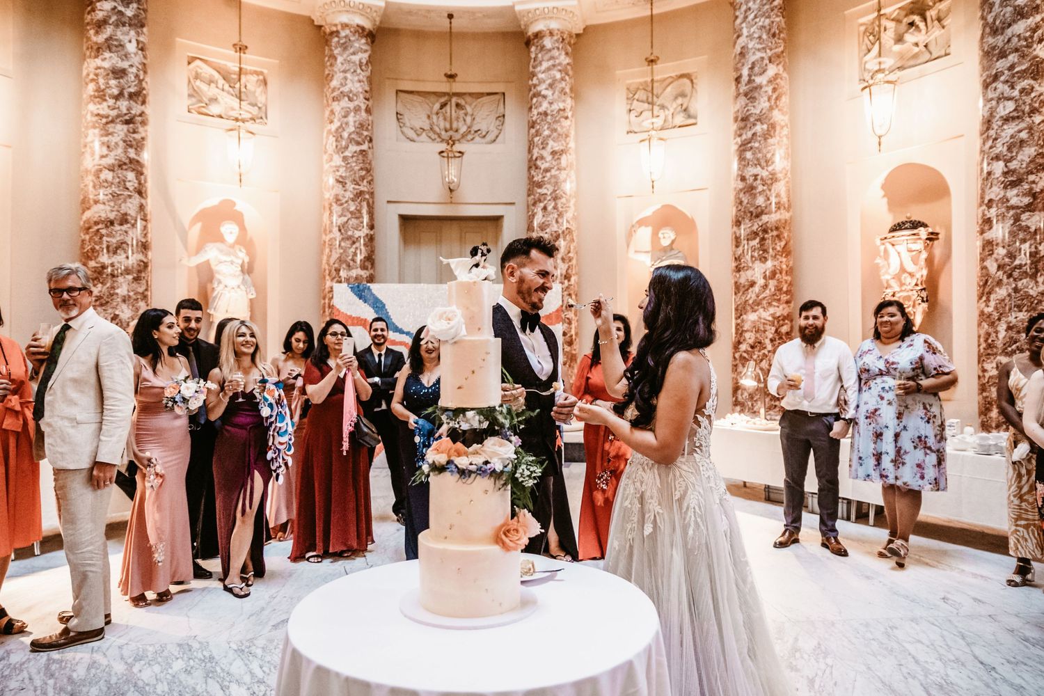 Wedding cake cutting ceremony in grand hall with ornate marble columns and gathered guests.