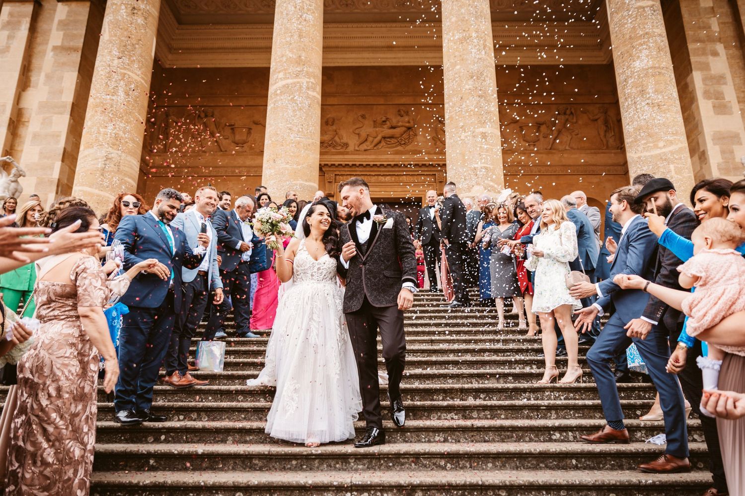 Wedding couple and guests celebrate with confetti on grand stone staircase.