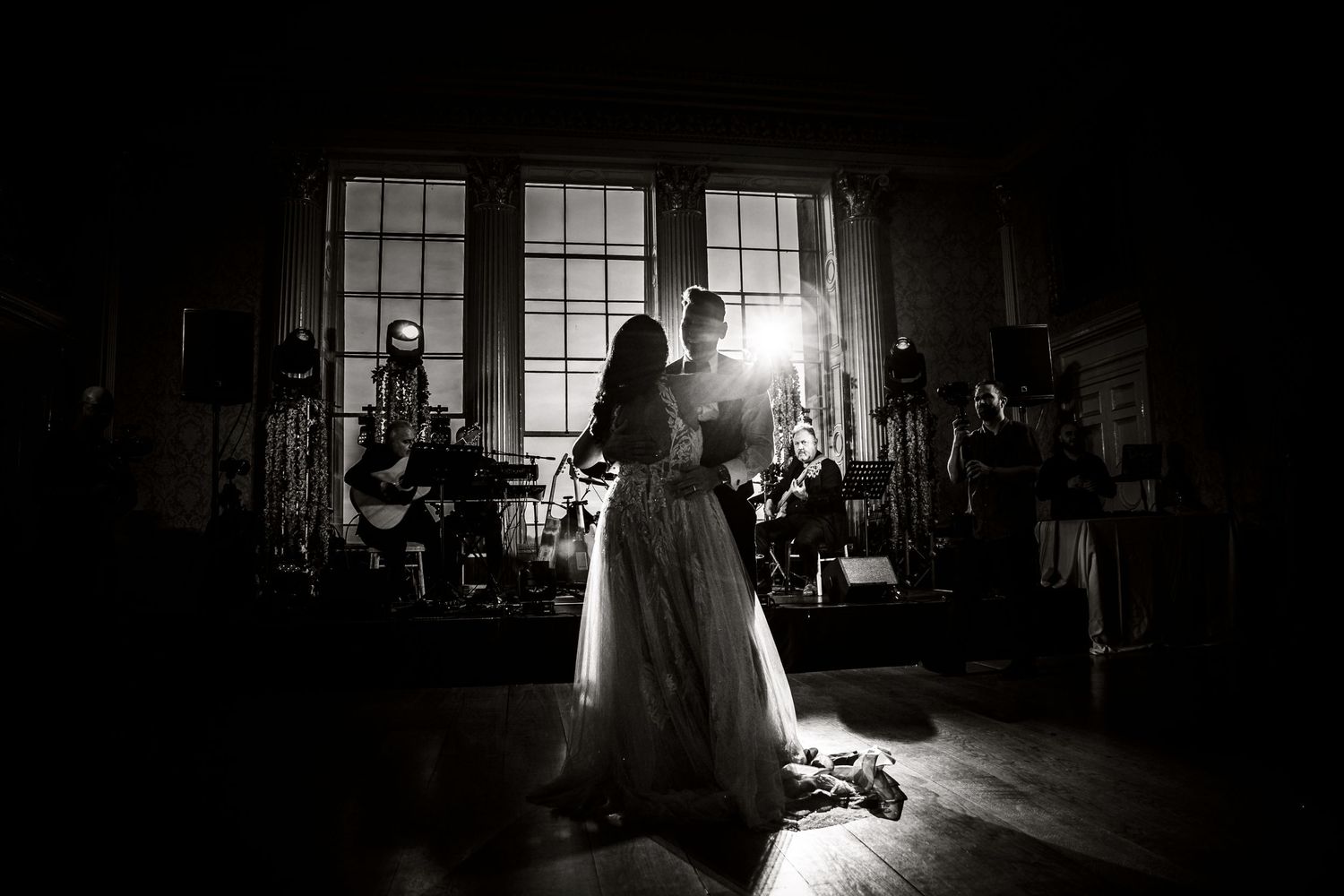 Black and white silhouette of dancing newlyweds with dramatic window lighting and band in background.