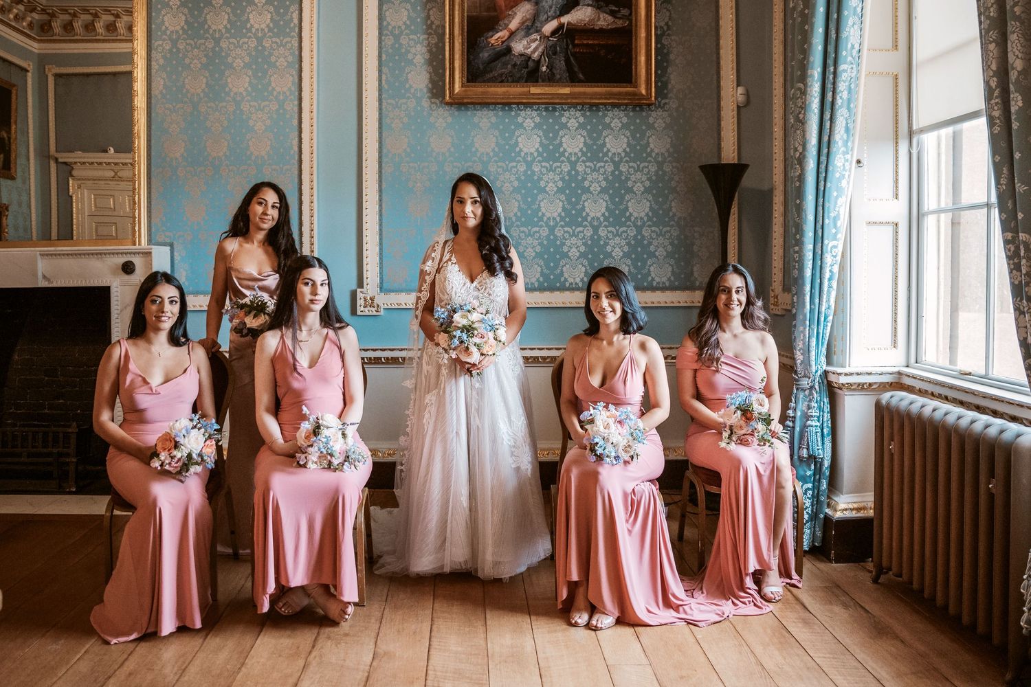 Wedding party poses in elegant blue room with bridesmaids in pink dresses.