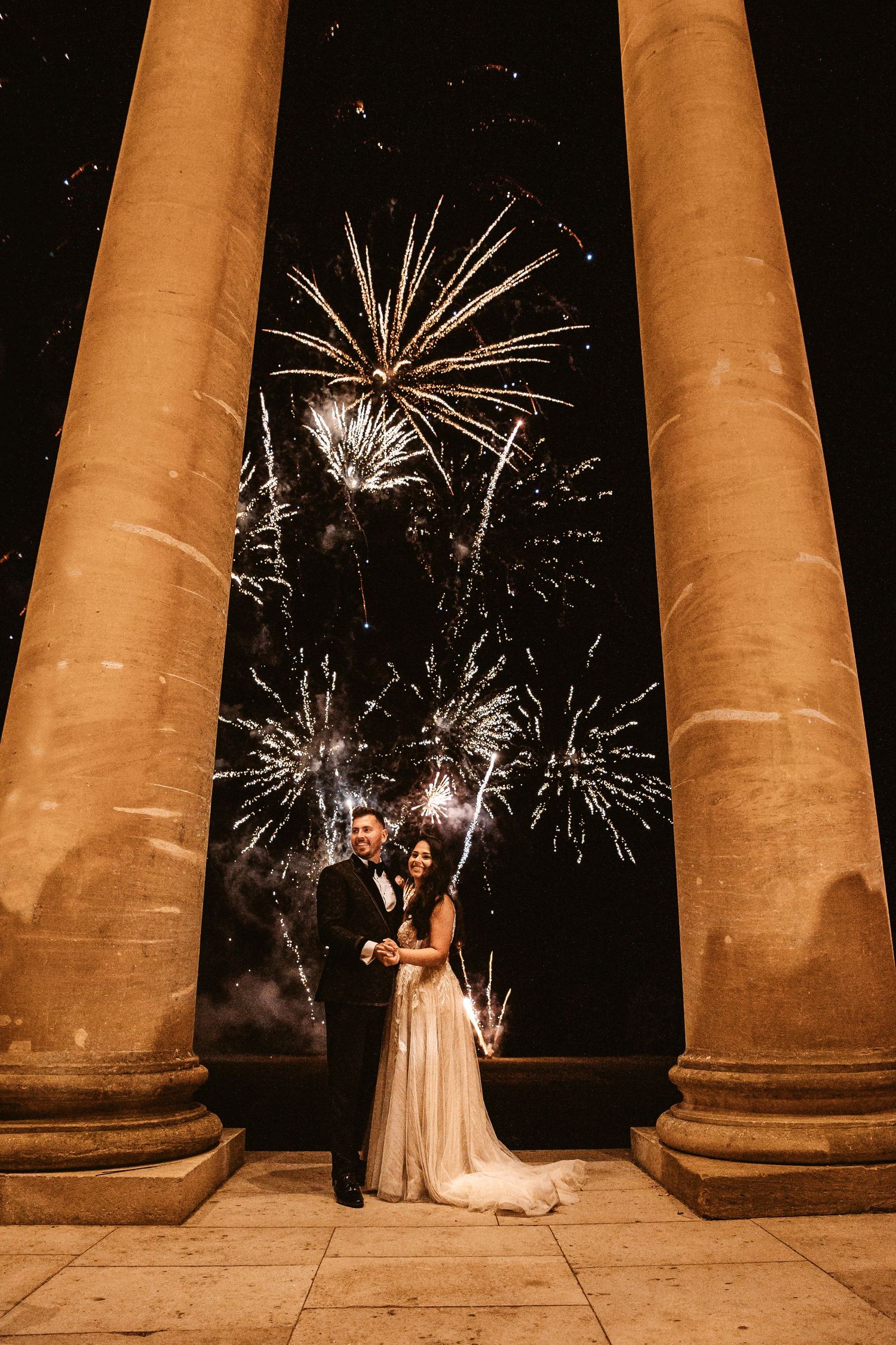 A spectacular fireworks display frames a couple between two majestic columns.