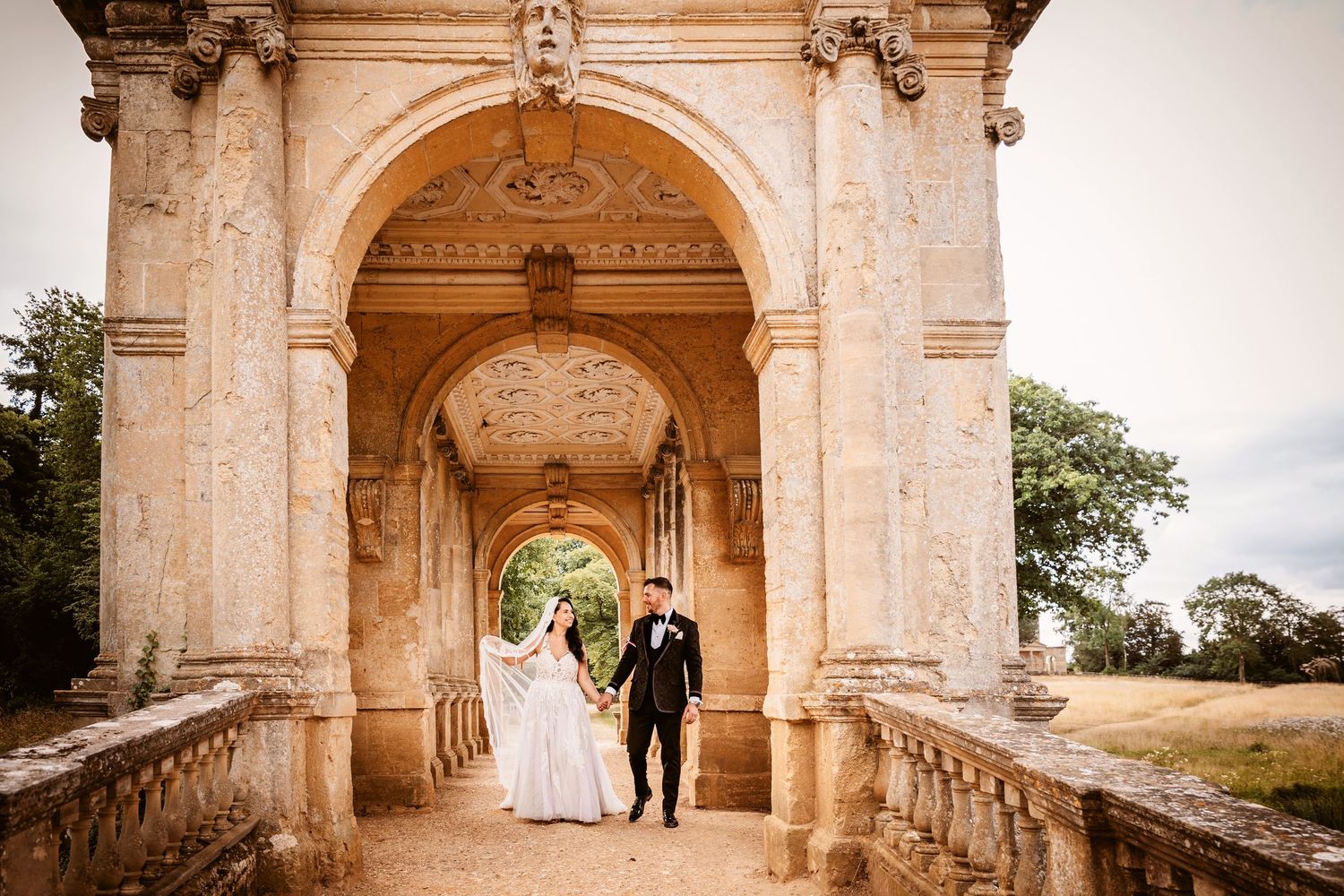 Couple stands under grand stone archway during wedding photo session.