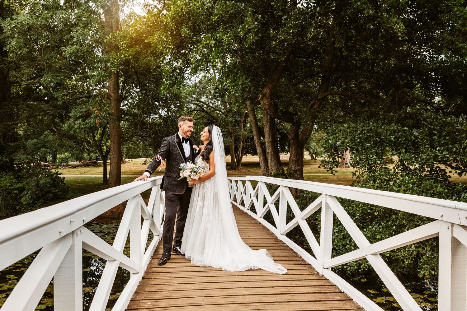 Newlyweds stand together on rustic white bridge during golden hour wedding photoshoot.