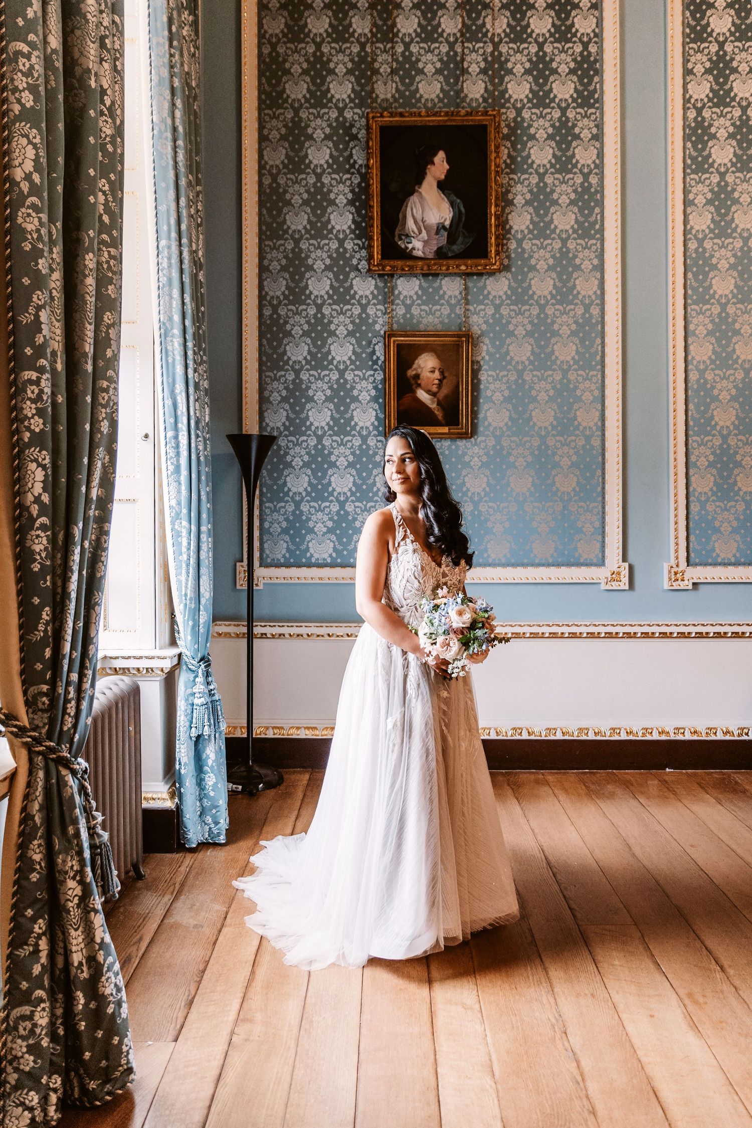 Bride in flowing white dress holds bouquet in elegant blue-wallpapered room with vintage portraits.