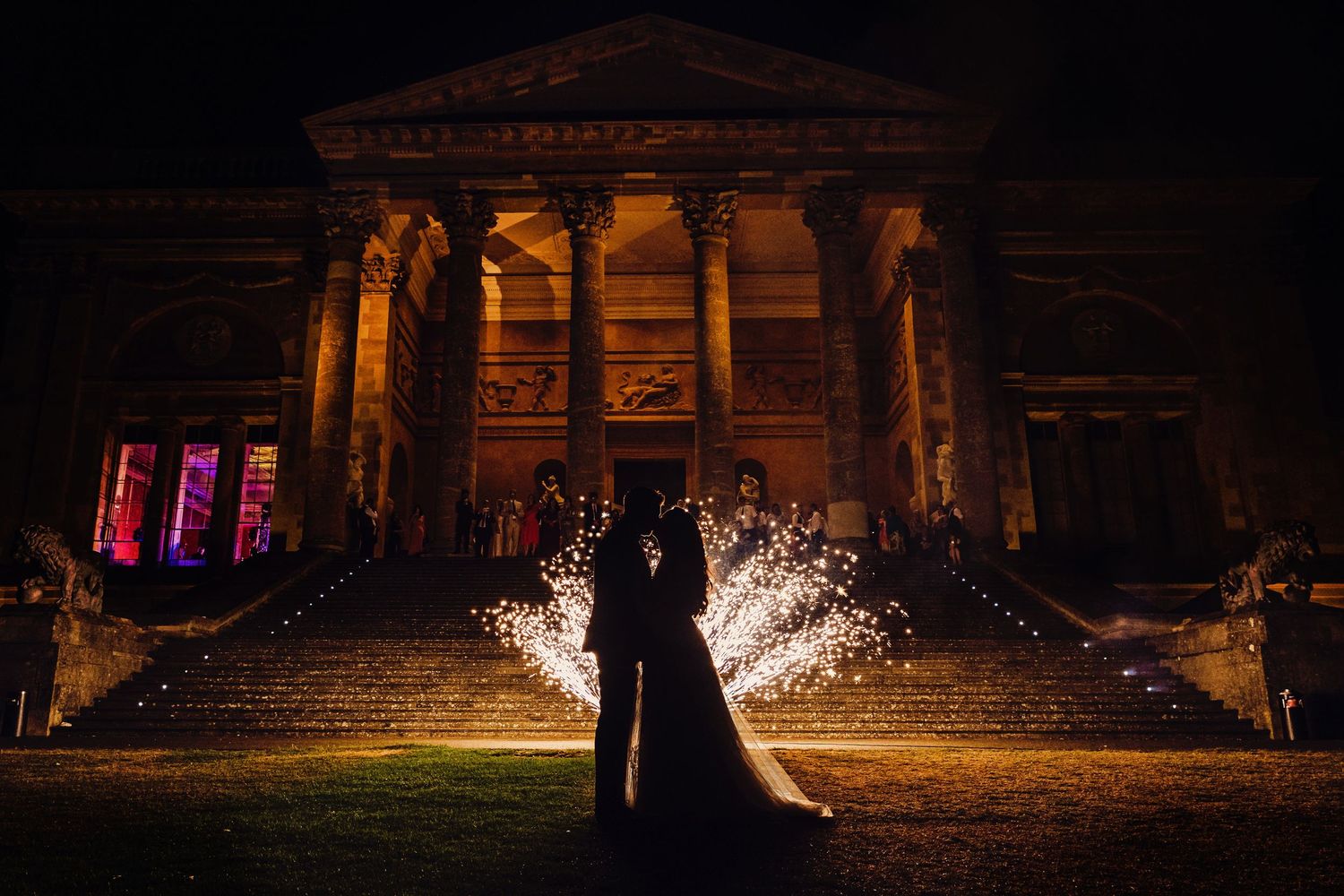 Dramatic sparkler fountains illuminate a couple in front of a classical building at night.