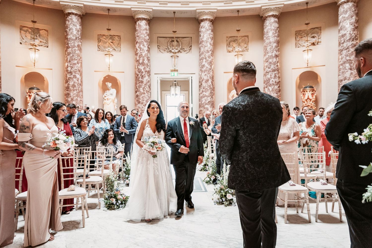 Wedding ceremony in progress inside ornate classical rotunda with marble columns and filled chairs.