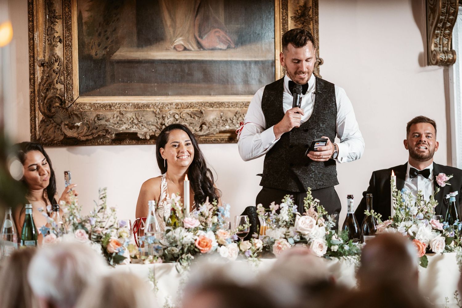 Wedding party seated at head table while speaker delivers toast in ornate historic venue.