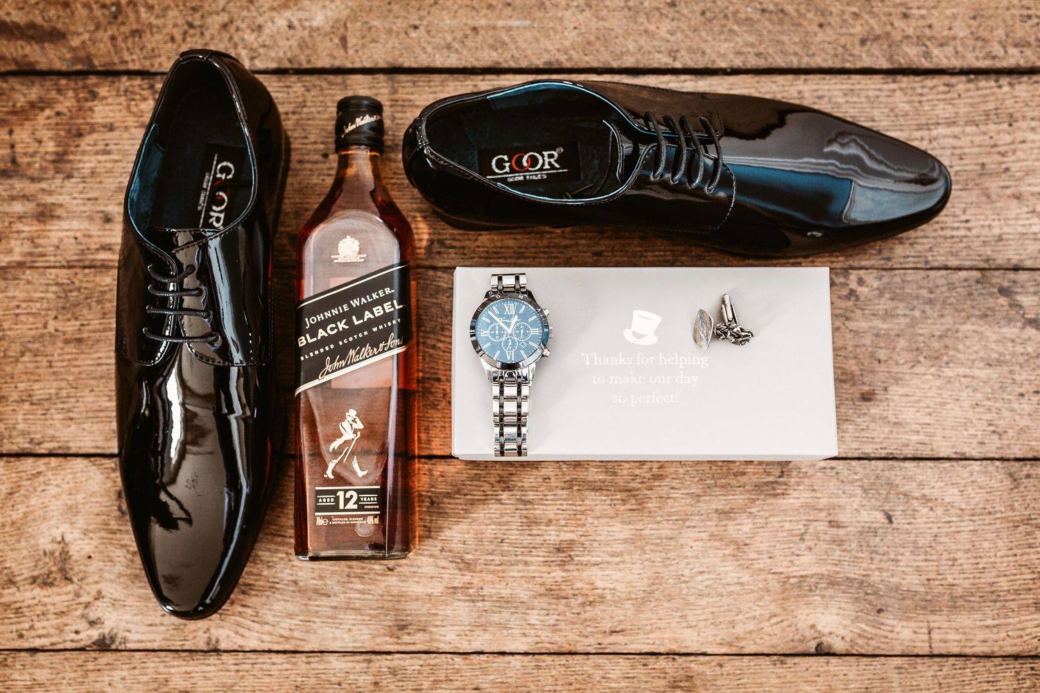Wedding accessories including dress shoes, Johnnie Walker whiskey, watch and cufflinks arranged on wooden surface.