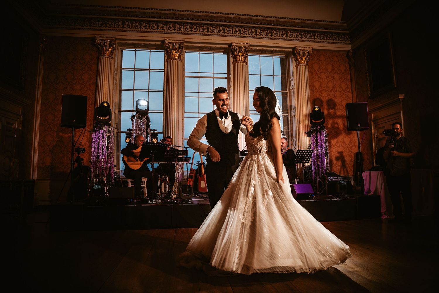 Wedding dance captured in silhouette against bright windows with live band performing on stage.