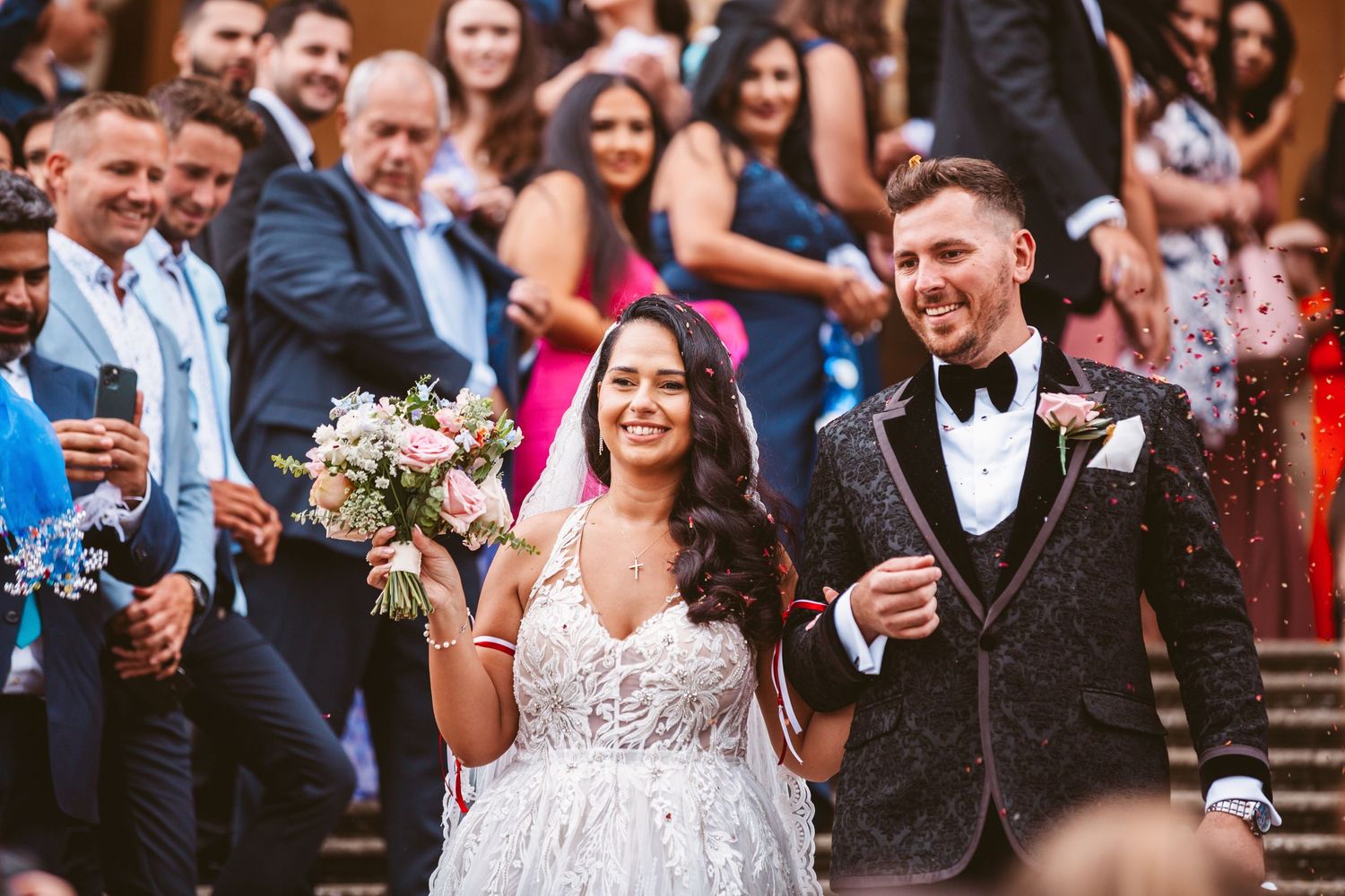 Joyful wedding couple walks down stone steps surrounded by cheering guests.