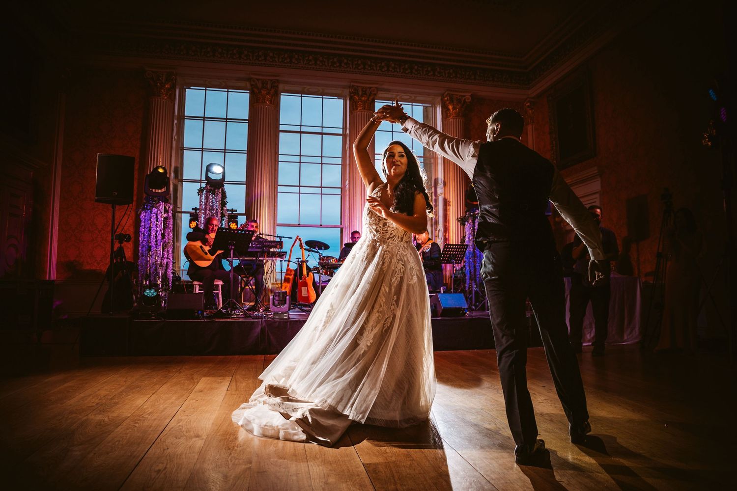 Dancing couple silhouetted against bright windows during wedding reception with live band.