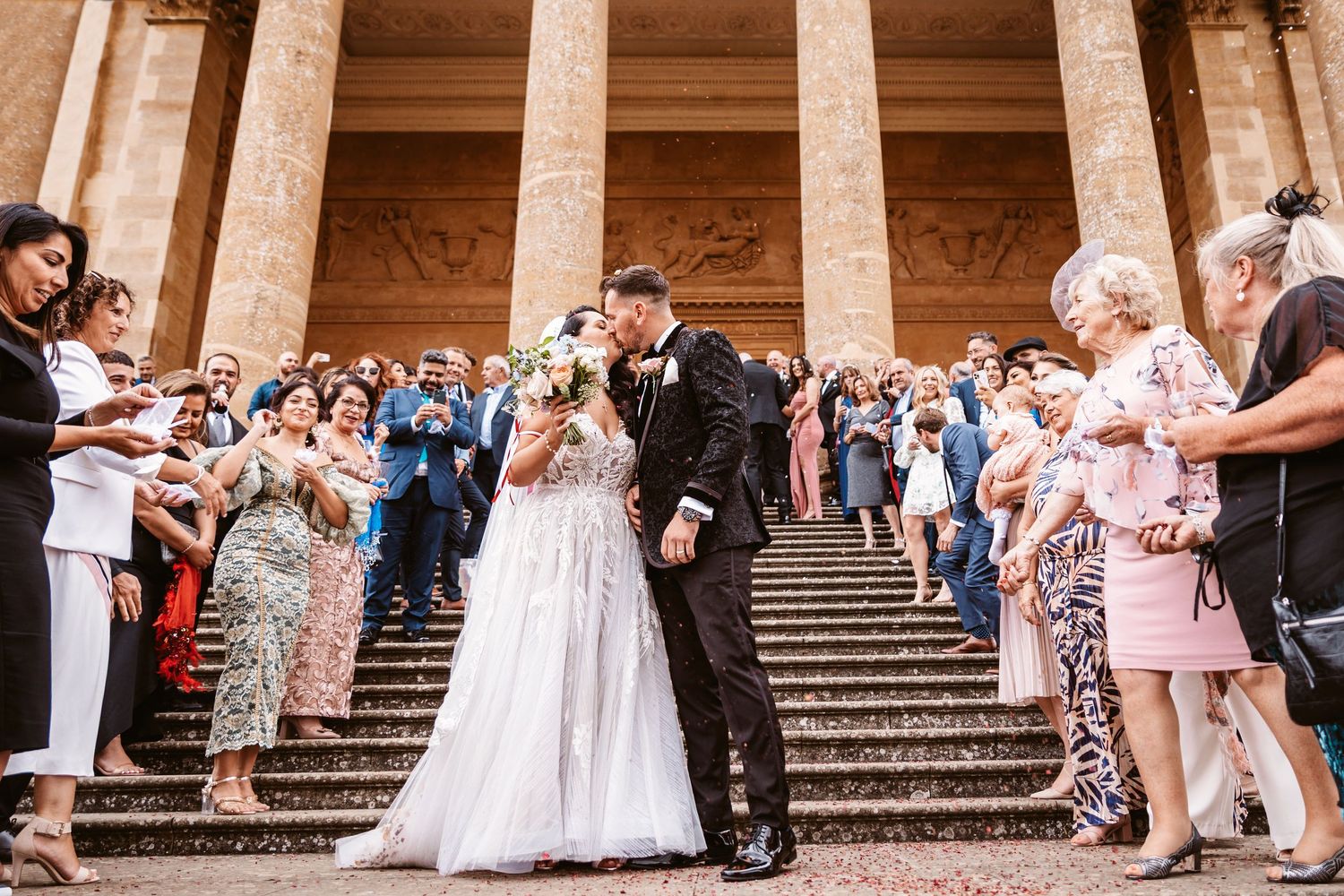 Wedding party exits while guests throw confetti on ornate building steps.