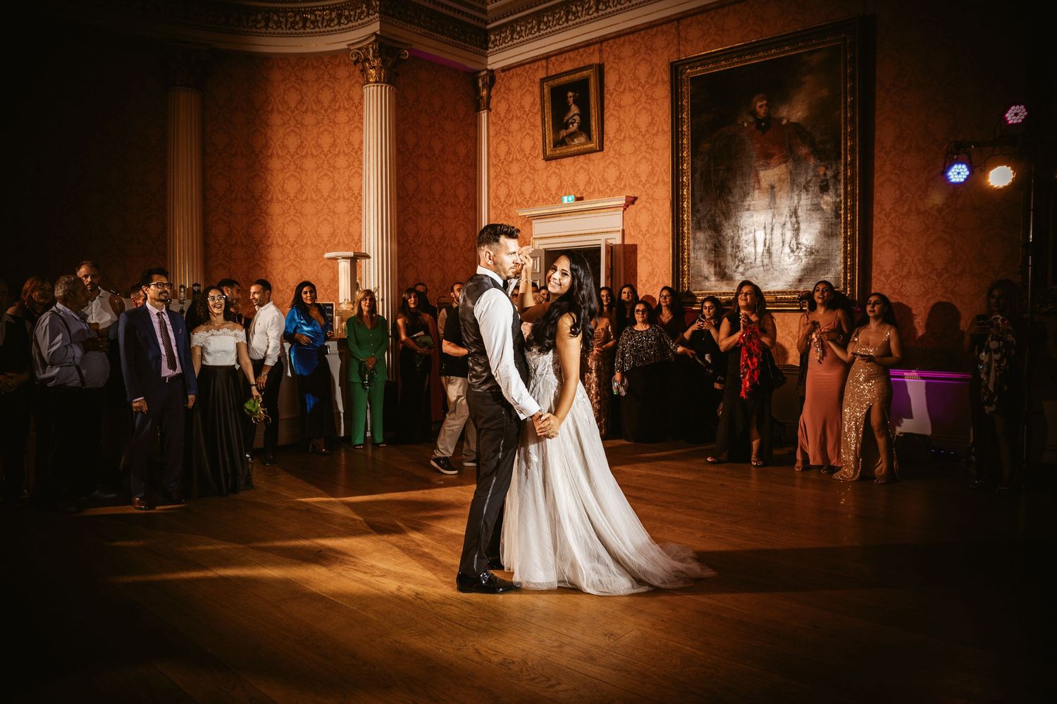Couple shares their first dance at an elegant wedding reception in a grand ballroom with ornate walls and lighting.