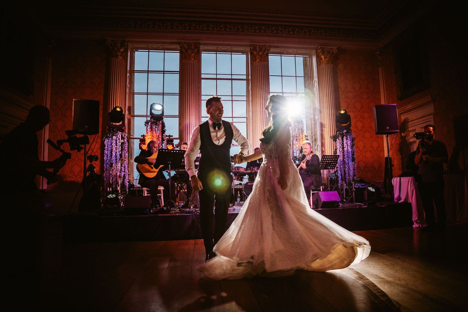 Bride's dress twirls during a romantic dance with dramatic backlighting from tall windows.