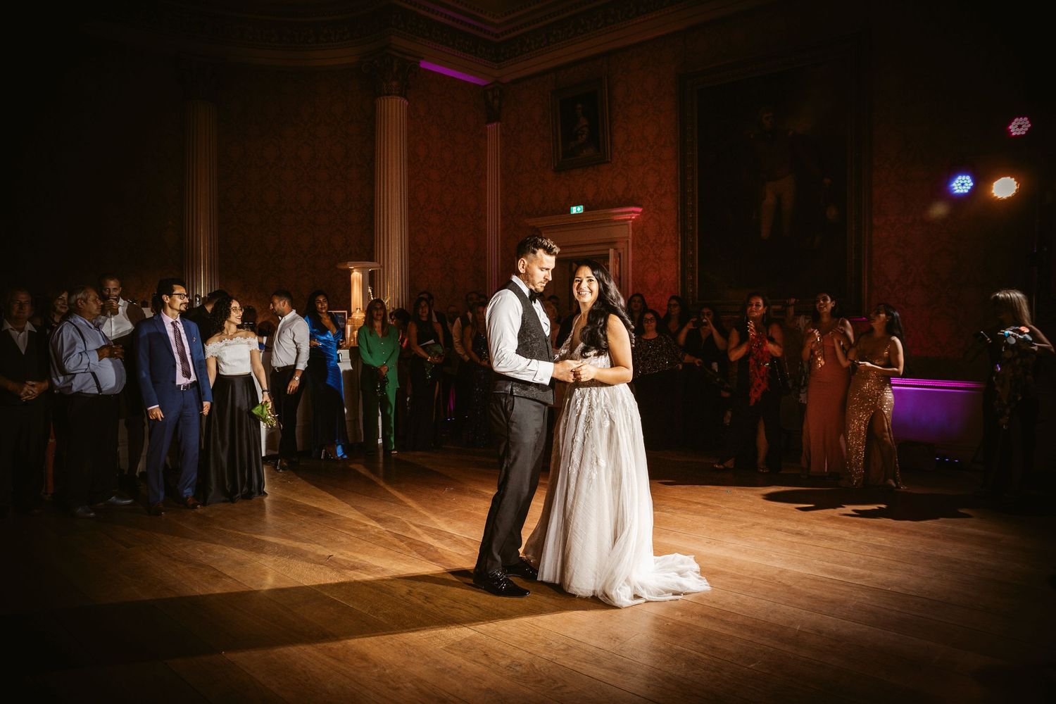 Wedding couple performs romantic first dance in dimly lit historic ballroom surrounded by guests.