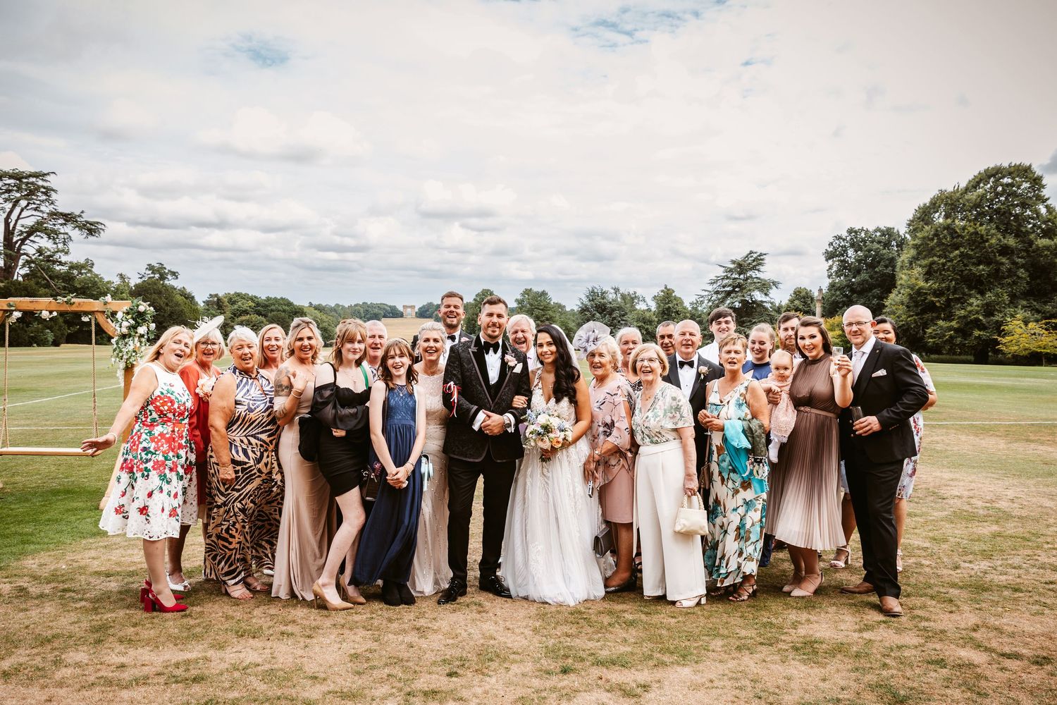 Large wedding party poses together on grassy field under cloudy sky during outdoor celebration.