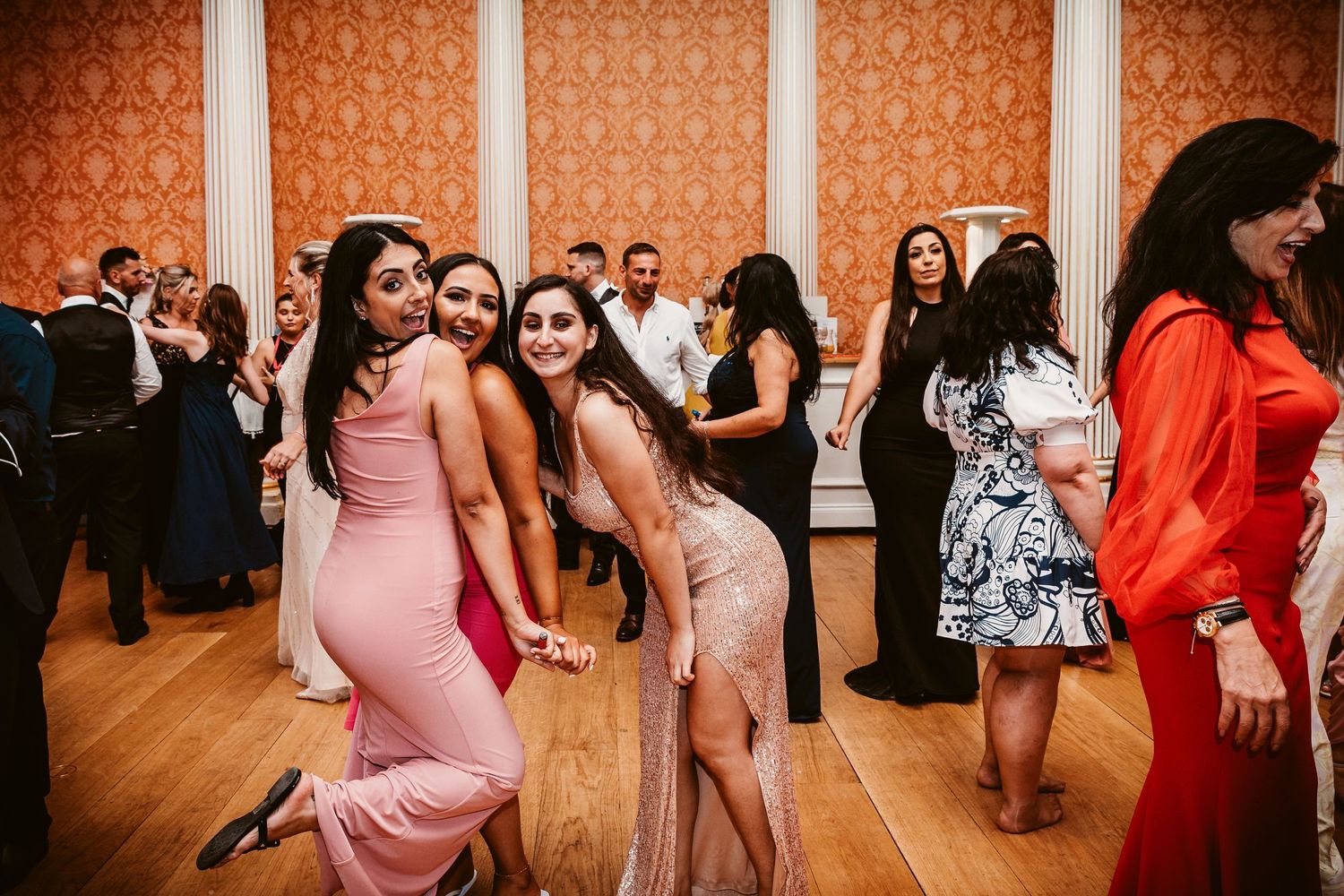 Wedding guests dance and celebrate on a dance floor with orange patterned wallpaper in the background.
