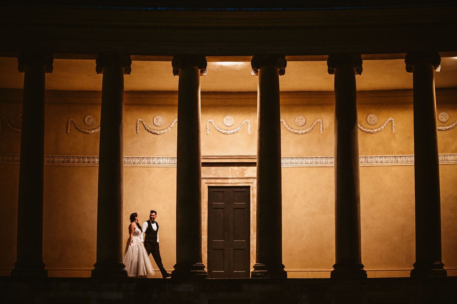 A romantic scene framed by classical columns against a warm-toned wall.