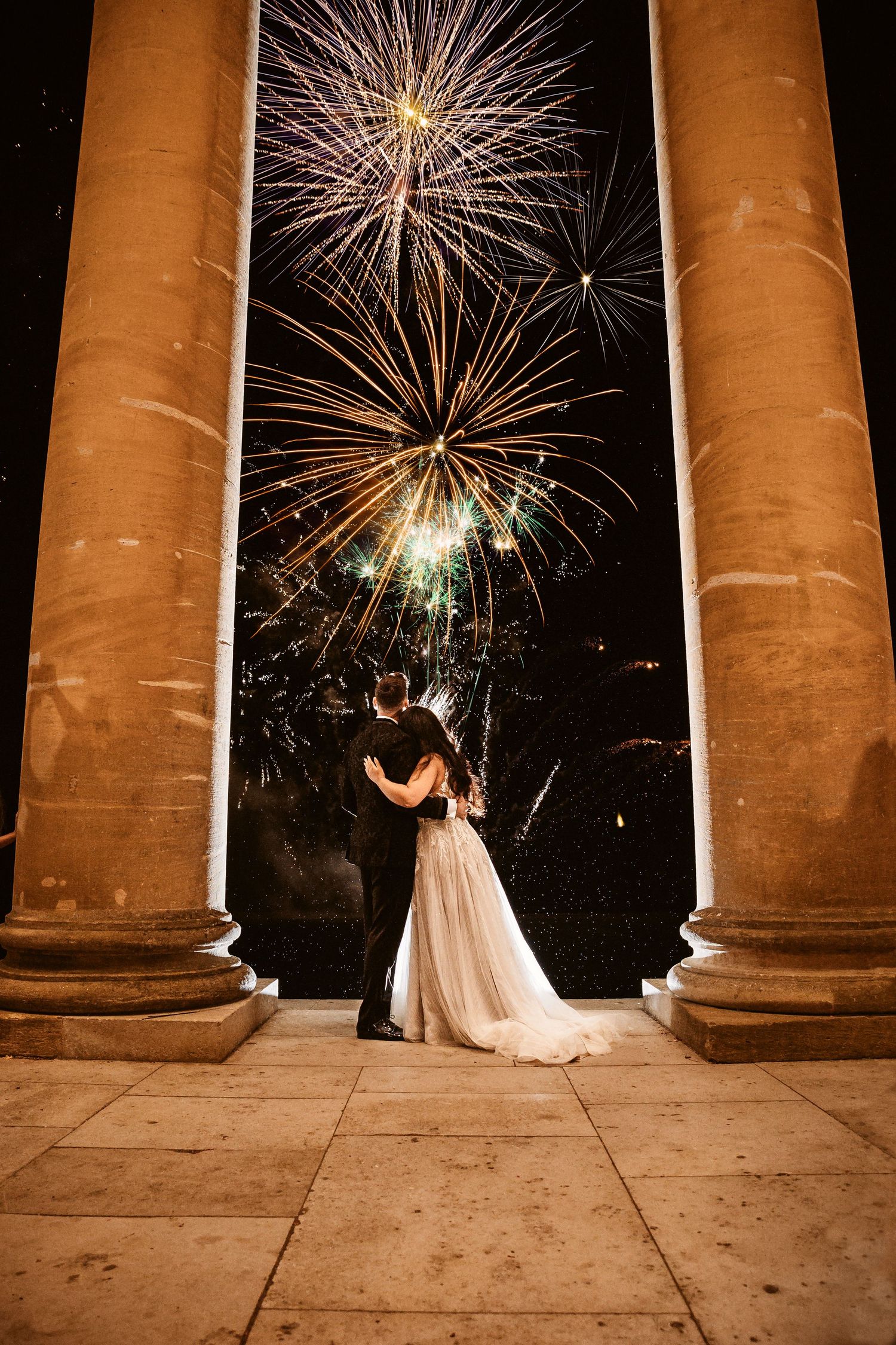 A romantic moment captured under classical columns with fireworks bursting in the night sky.
