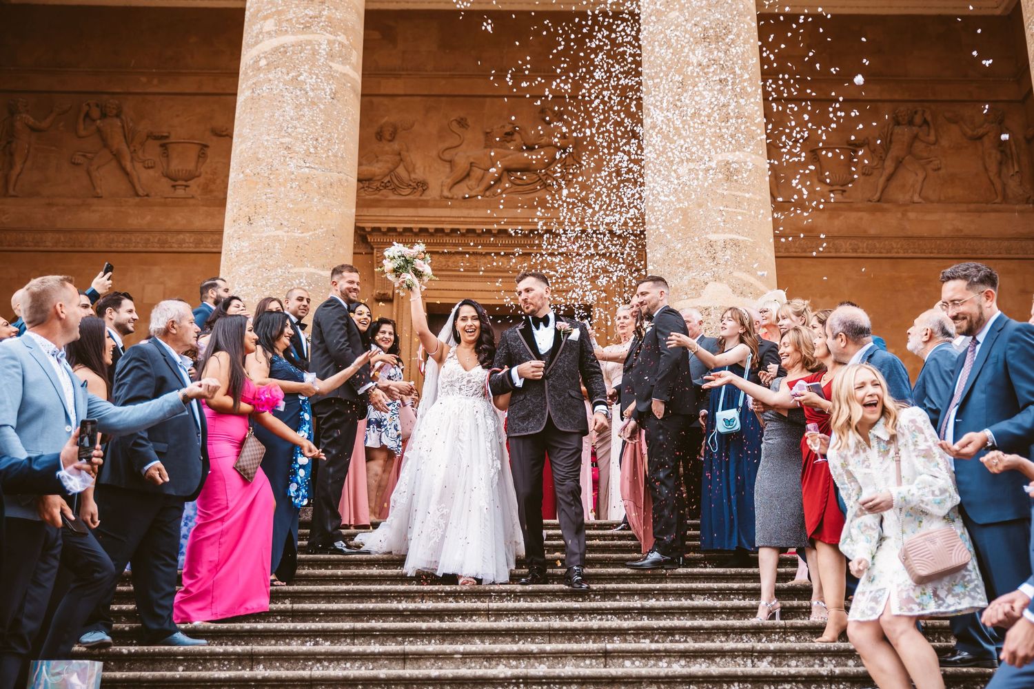Wedding guests shower confetti over the newlyweds on grand stone steps.