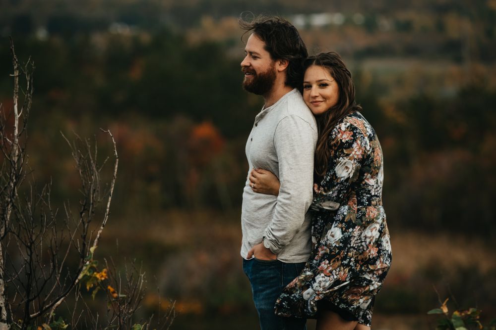 Couple in casual attire standing together on hilltop with fall foliage in background.