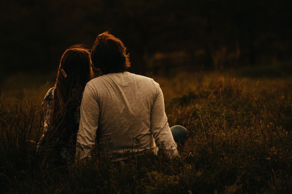 Couple sits together on grassy slope during sunset engagement session.