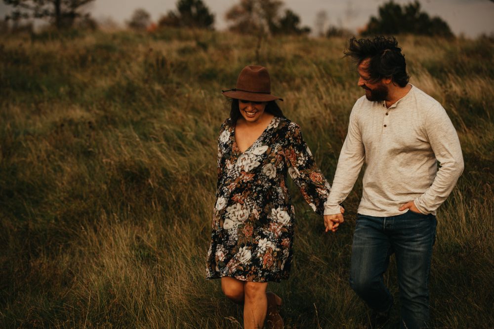 Couple walks hand in hand through tall grass at sunset.