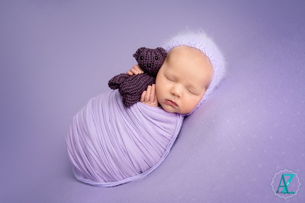 Newborn photography on purple backdrop with lavender wrap and dark bow accent.