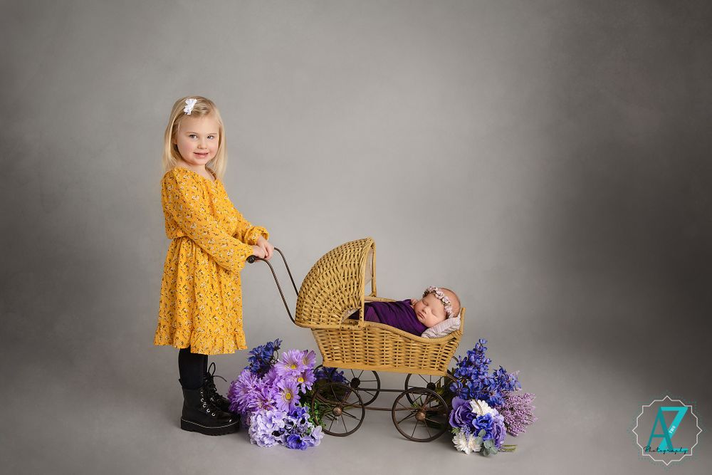 Studio portrait with vintage wicker pram, yellow dress, and purple flower decorations.