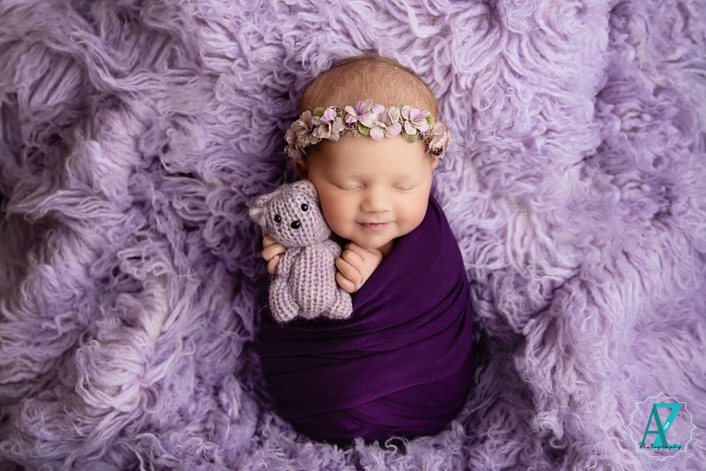 Newborn wrapped in purple fabric with floral headband lying on purple fur backdrop with stuffed toy.