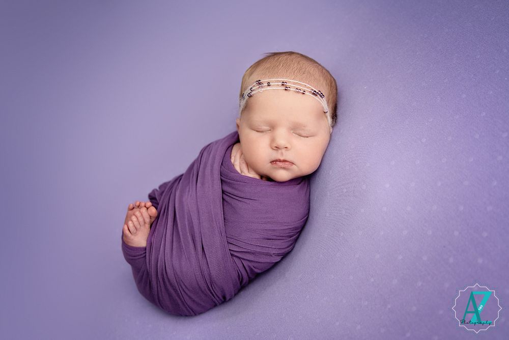 Sleeping newborn wrapped in purple fabric with delicate headband against purple background.