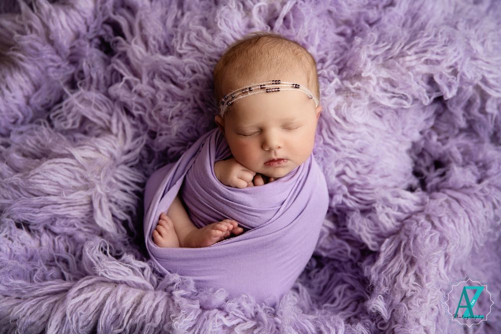 Peaceful newborn in purple wrap on fluffy purple blanket wearing delicate headband.
