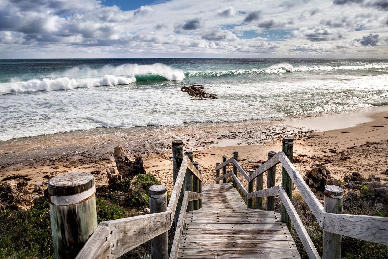A wooden staircase down the beach with a massive wave crushing down on a sunny day