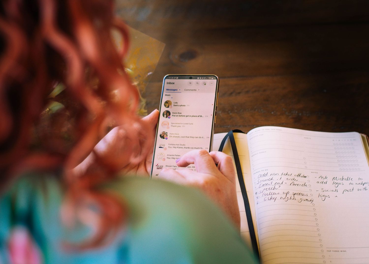 Student taking notes on smartphone next to open notebook on wooden desk with soft focus red hair.
