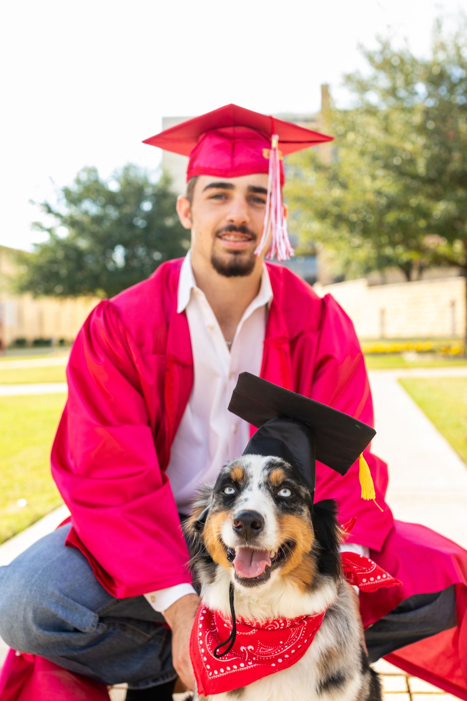 Male graduate in red cap and gown poses with Australian Shepherd dog wearing graduation cap.