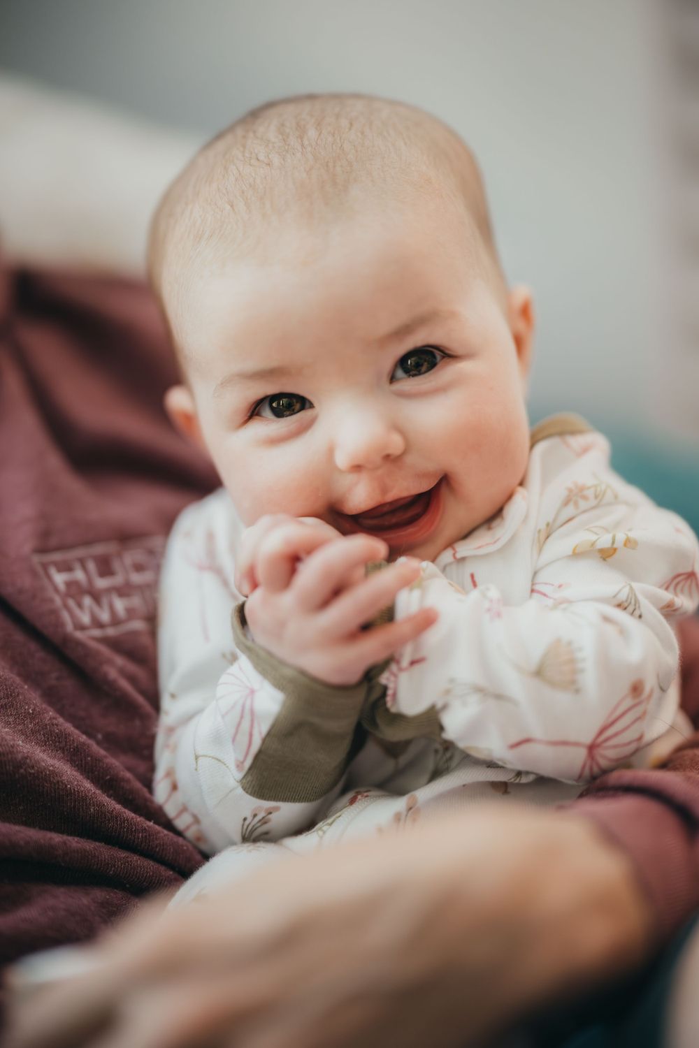 Adorable baby in white clothing smiles while lying on brown blanket.