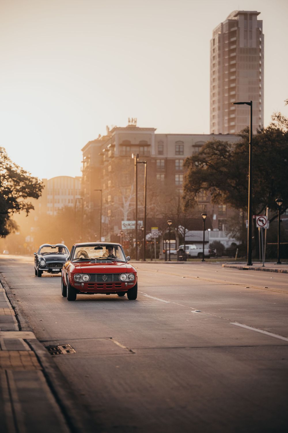Urban street scene at sunset with classic cars and city buildings in the background.