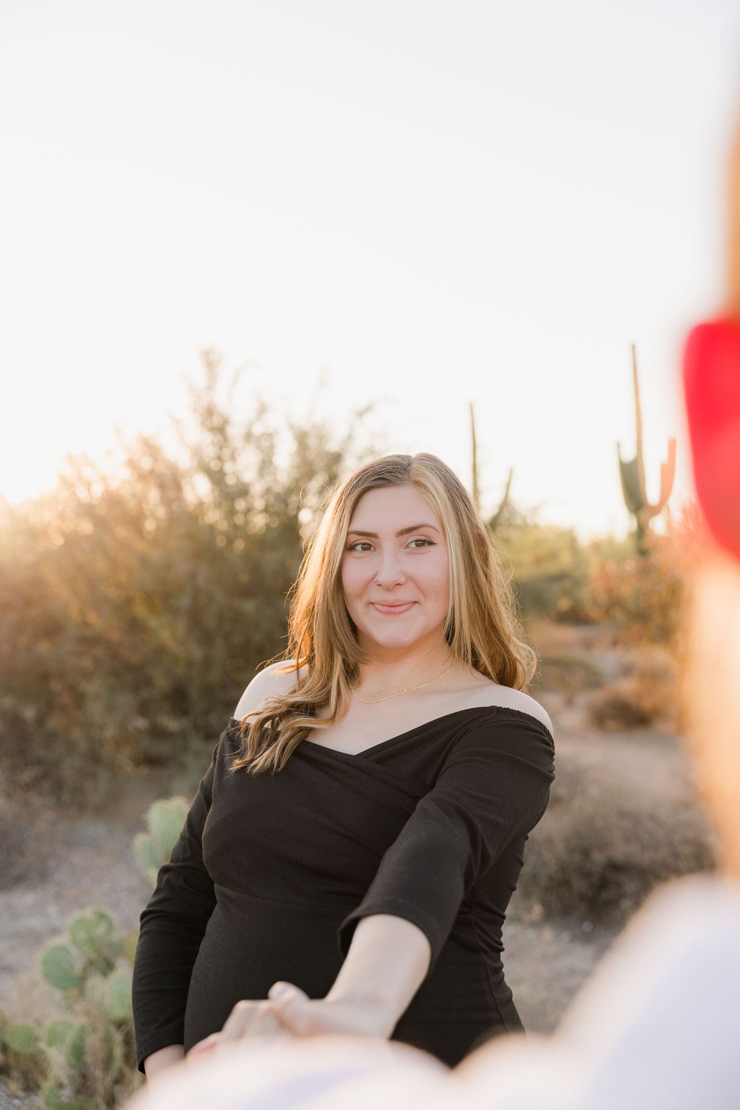 Person in black off-shoulder dress poses in desert landscape at sunset.