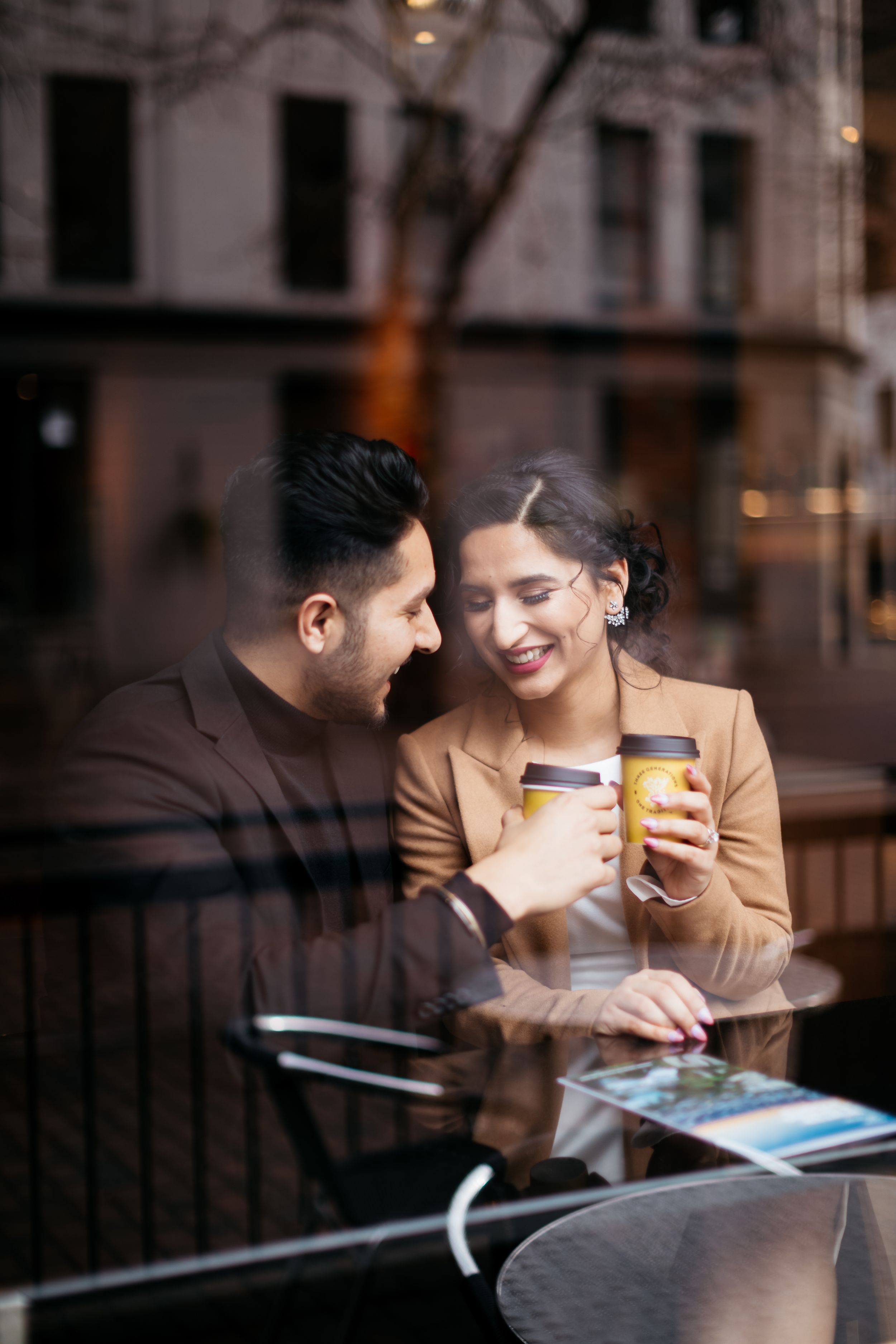 A couple shares coffee and conversation at a cafe table seen through window glass.