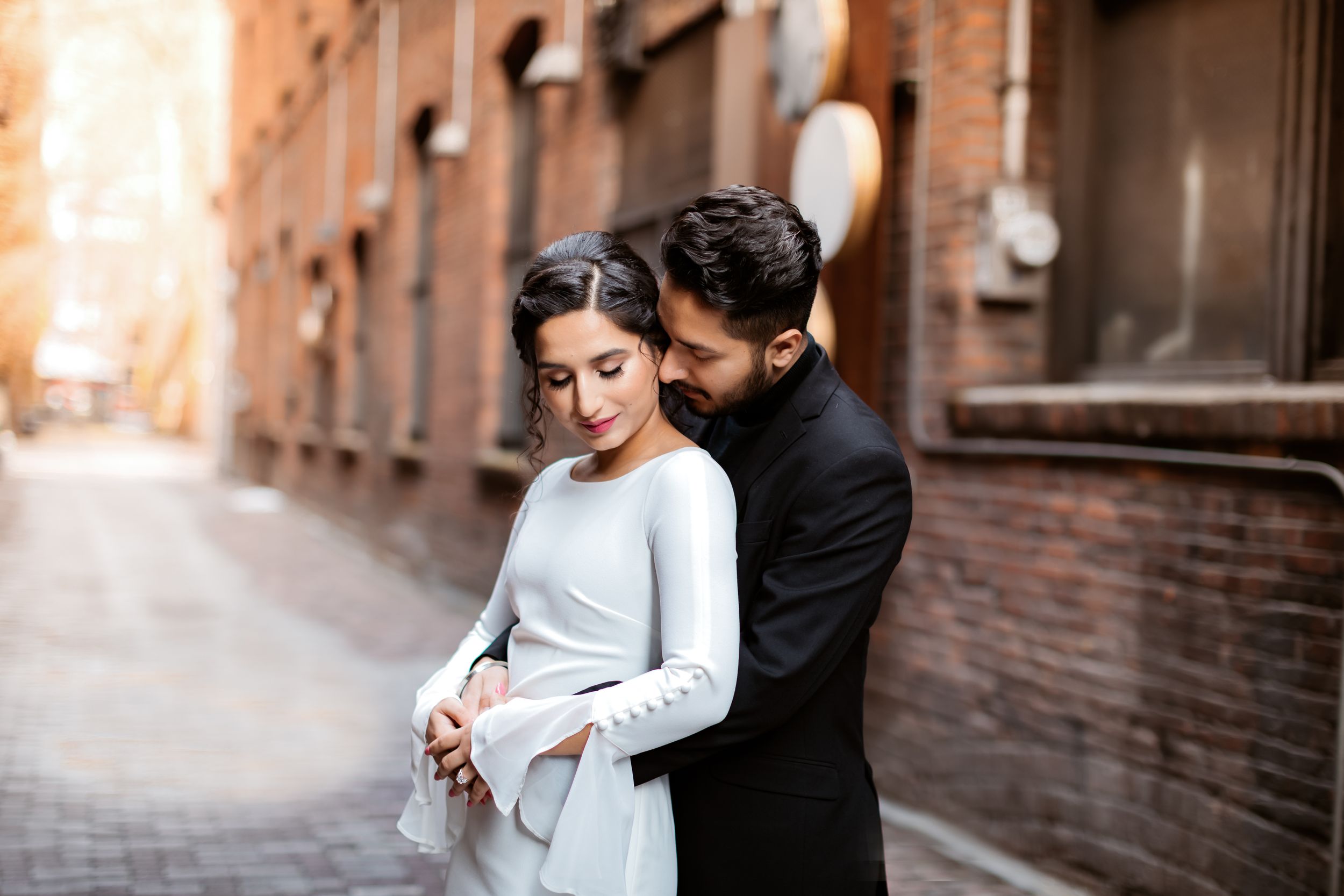A couple shares an intimate embrace in an urban alleyway with brick buildings.