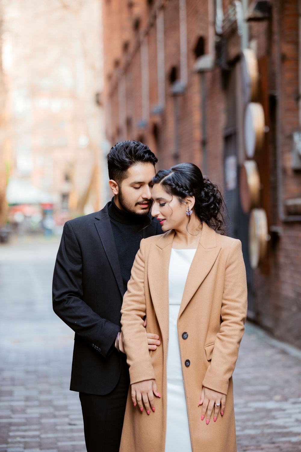 A romantic couple in stylish outfits embrace on a brick-lined street, with her wearing a camel coat and him in black.