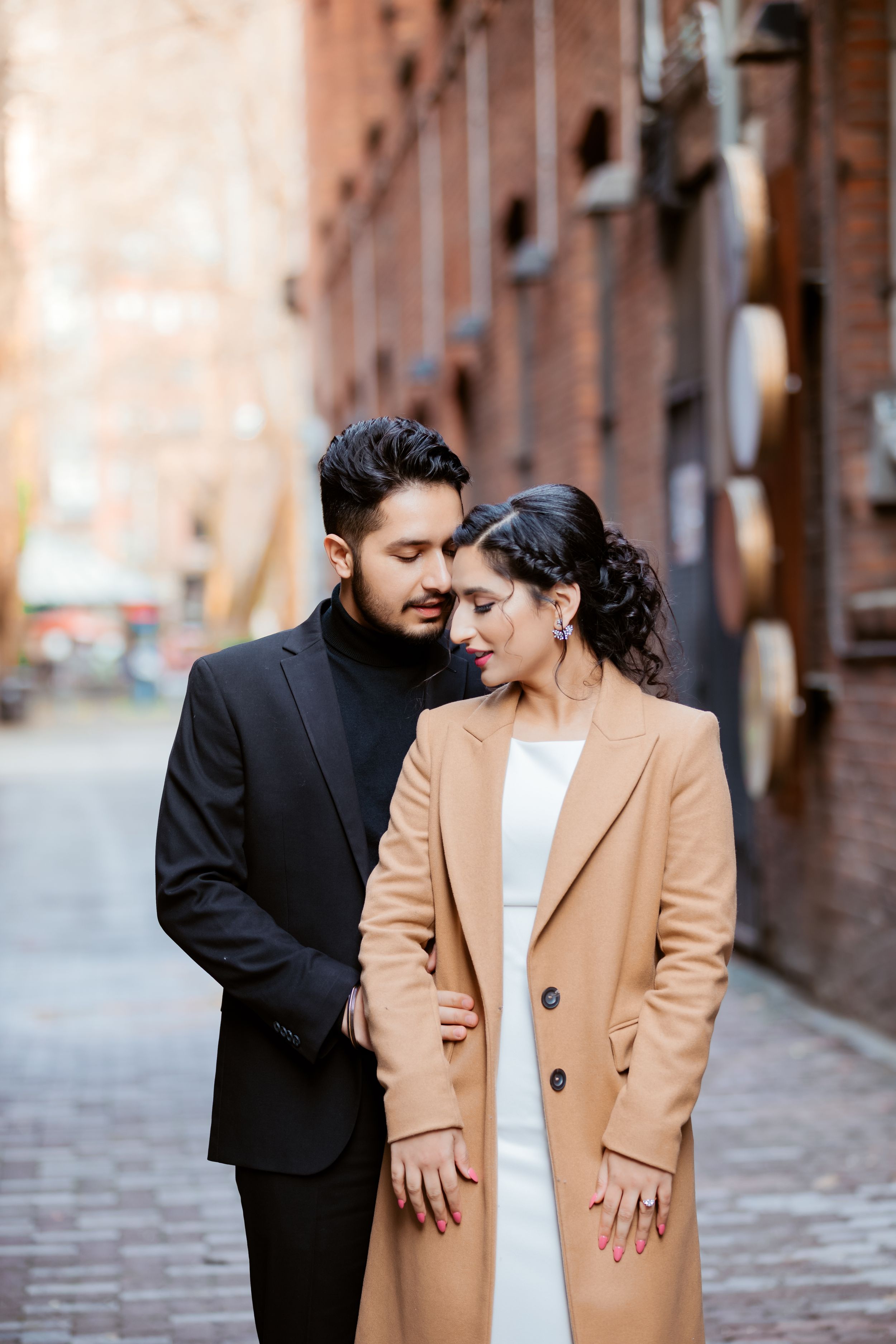 A romantic couple in stylish outfits embrace on a brick-lined street, with her wearing a camel coat and him in black.