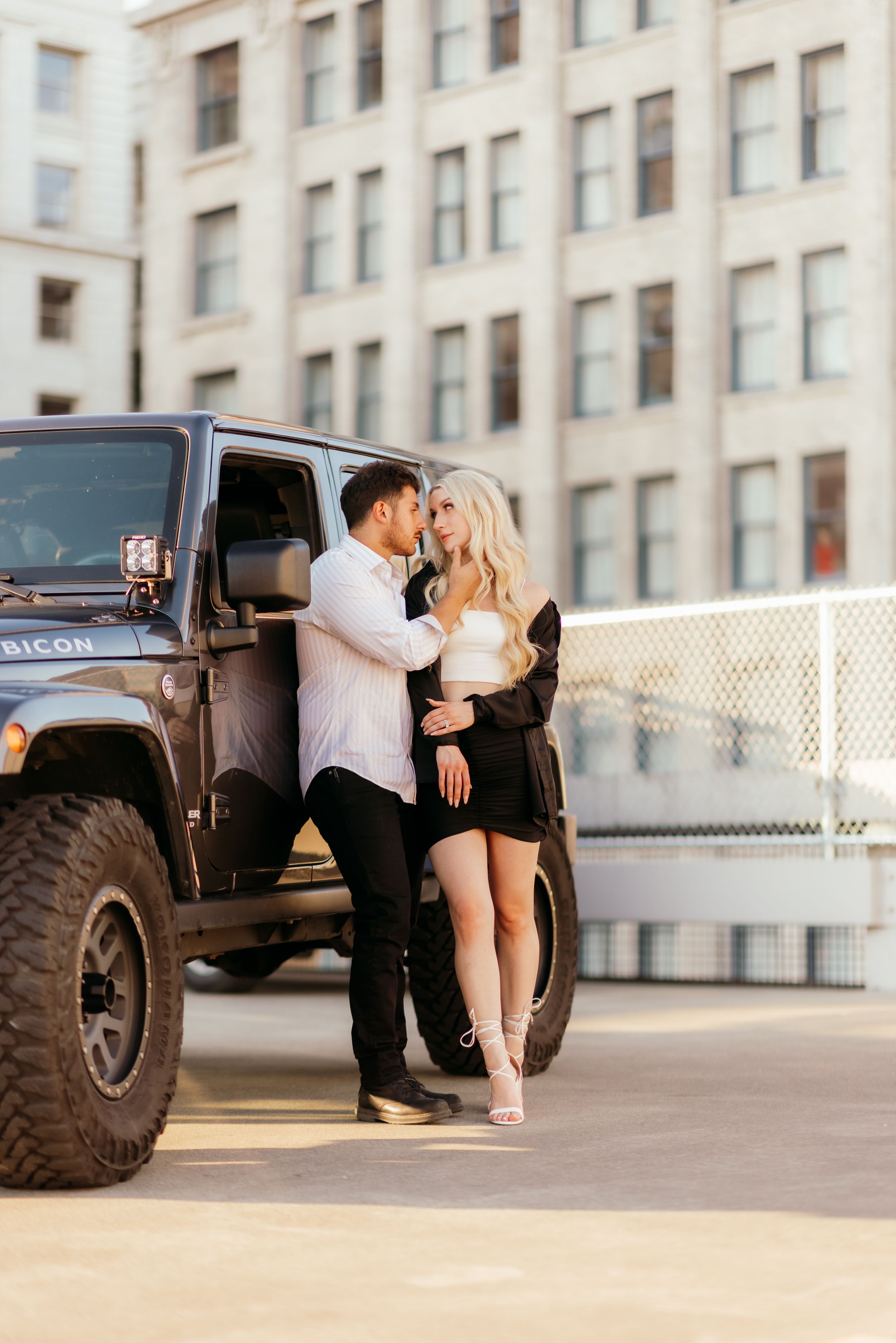 A couple shares an intimate moment beside a black Jeep Wrangler in an urban setting.