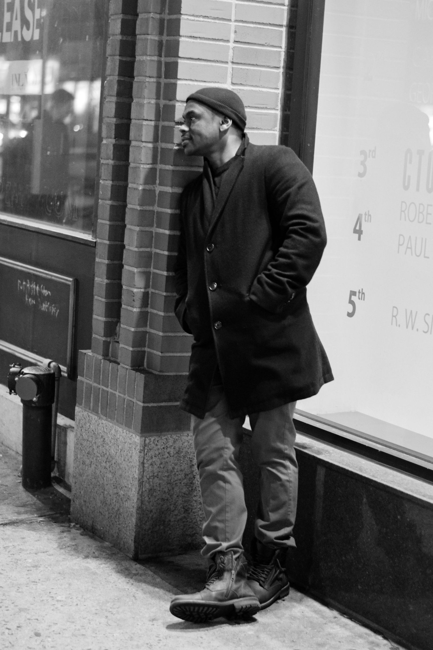A man in winter coat and cap leans against a brick building wall in an urban black and white scene.