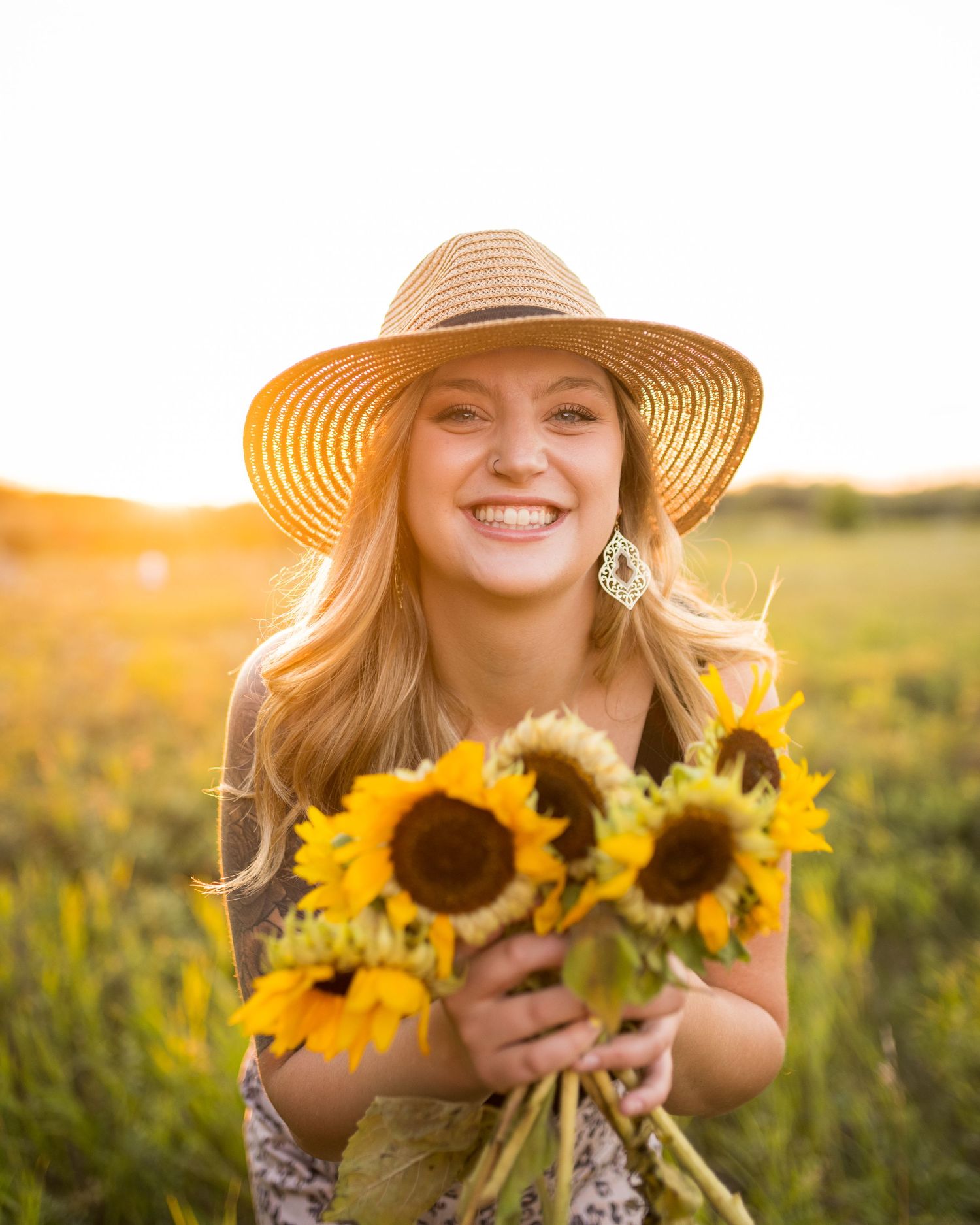 Happy person holding sunflowers in a field during golden hour sunset while wearing a straw hat.