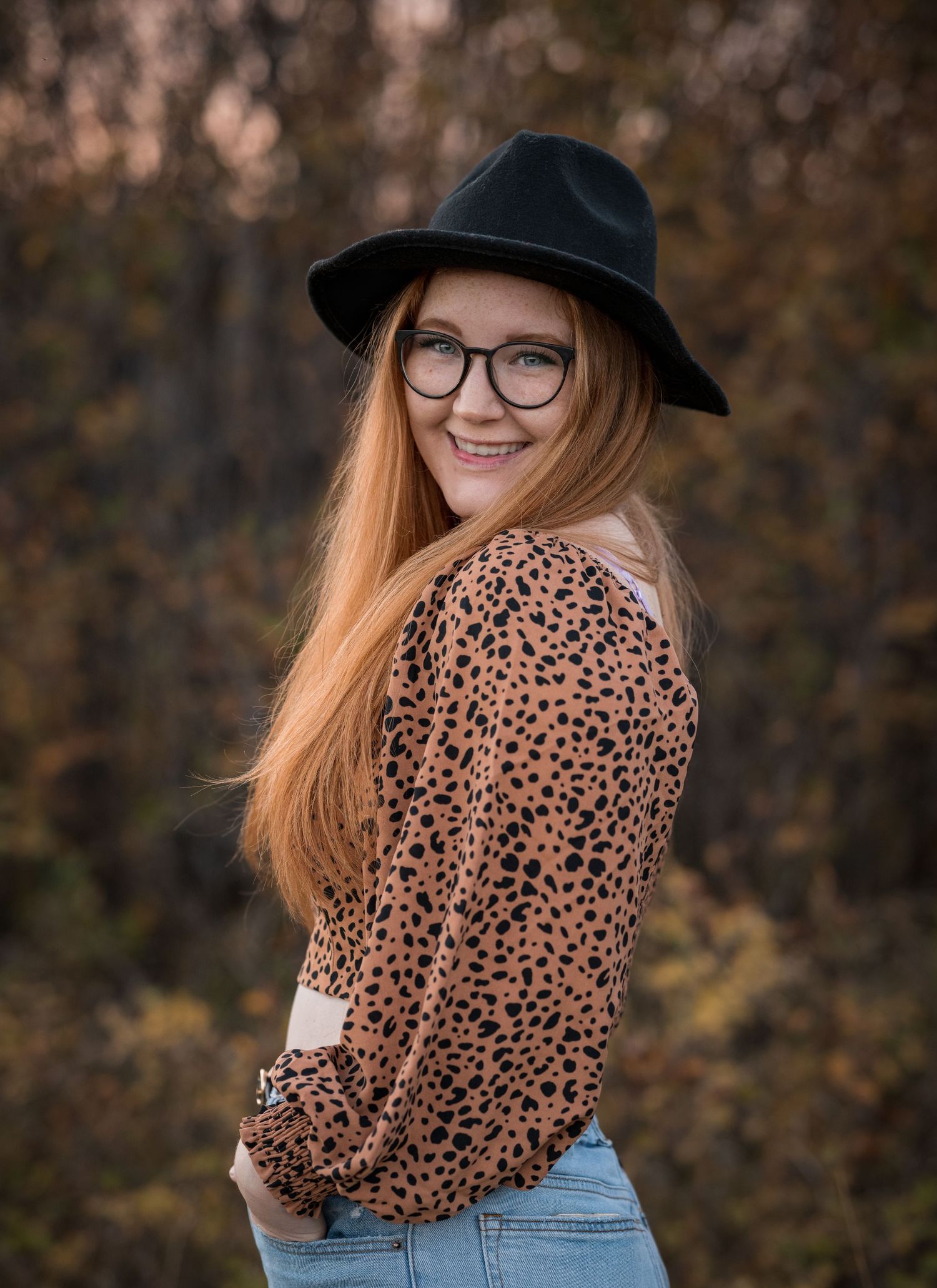 Portrait of someone in leopard print and black hat against autumn background.