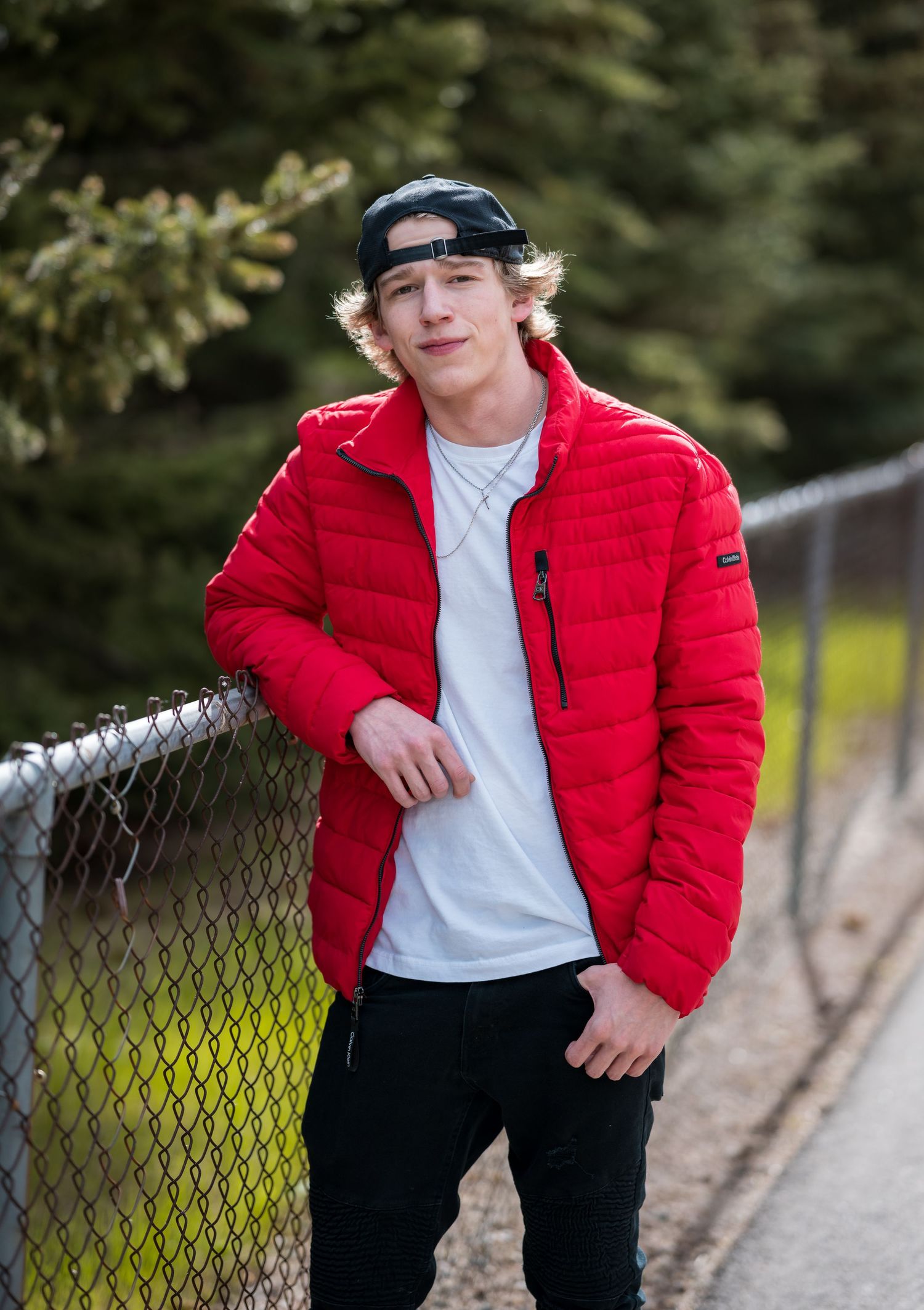 Person in red puffy jacket and baseball cap poses casually against chain link fence outdoors.